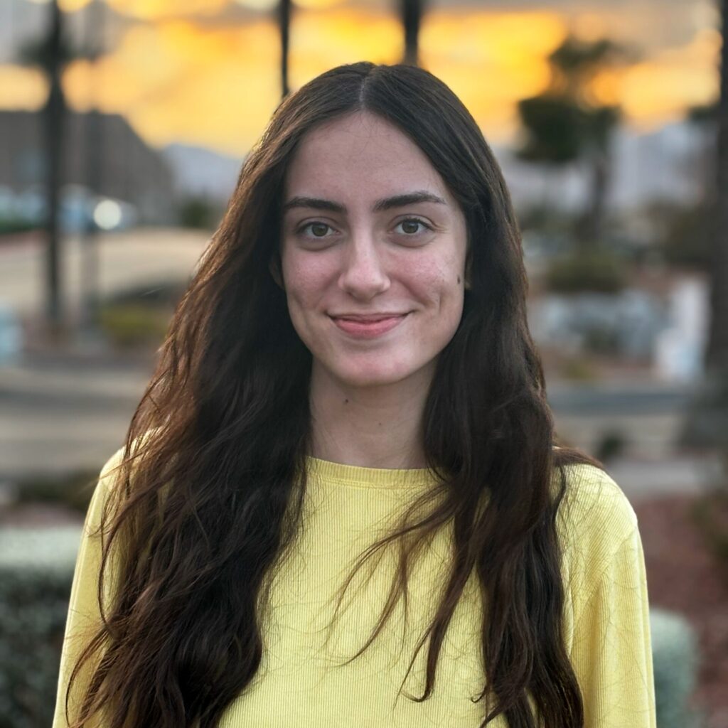 Young woman wearing a yellow top, with long hair. She is looking straight to the camera and smiling. 
