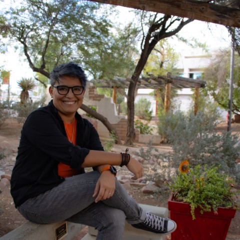 Mujer con lentes de marco azul, orange t-shirt and black shirt. She is smiling and locking at the camera and is sitting in the top of a concreate bench.